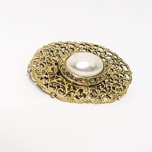 Puede incluir: Broche ovalado dorado con un gran centro de perla blanca redonda. El broche presenta un diseño de filigrana calada ornamental alrededor de la perla. Una pieza de joyería vintage clásica.