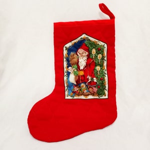 May include: Red quilted Christmas stocking with a festive scene of Santa Claus holding a bag of gifts and a Christmas tree with ornaments. The stocking has a white trim and a loop at the top.