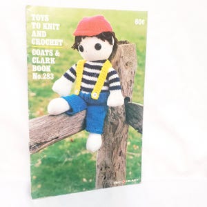 May include: A vintage book cover titled "Toys to Knit and Crochet" featuring a knitted doll wearing a red cap, striped shirt, blue pants, and yellow suspenders. The doll is sitting on a wooden fence. The book is from Coats & Clark.