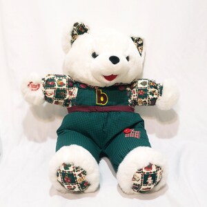 May include: A white plush teddy bear wearing green overalls with a red and green patterned shirt and accents. The overalls have a large yellow "b" on the front. The bear has a red mouth and black eyes. The bear's paws and ears have a matching pattern.