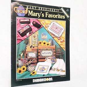 May include: A book cover titled "Mary's Favorites" by Mary Engelbreit. The cover features various cross-stitch designs, including a teapot, floral patterns, and the word "WELCOME". The book is from Dimensions.