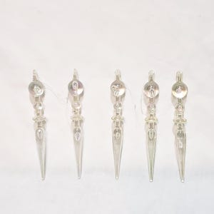 May include: Five clear glass icicle ornaments with a silver iridescent finish. Each ornament has a round bead at the top and a pointed tip.