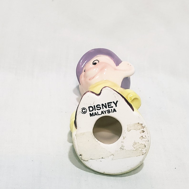 May include: A ceramic figurine of a dwarf from the Disney movie Snow White and the Seven Dwarfs. The dwarf is wearing a yellow shirt and a purple hat. The figurine is made in Malaysia and has the Disney copyright symbol on the bottom.