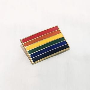 Puede incluir: Un pin rectangular con el diseño de la bandera del arcoíris, con franjas rojas, naranjas, amarillas, verdes, azules y moradas. El pin tiene un borde dorado y está sobre un fondo blanco.