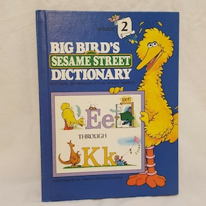 May include: A blue book with the title "Big Bird's Sesame Street Dictionary" featuring Jim Henson's Sesame Street Muppets. The book cover features Big Bird holding a sign with the letters "Ee" and "Kk" and the words "Through" and "Exit".