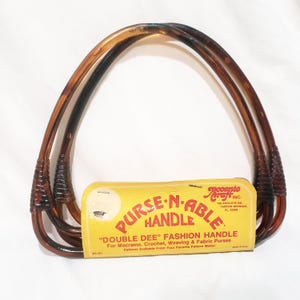 May include: Two brown plastic purse handles with a yellow and red label that reads "PURSE-N-ABLE HANDLE" and "DOUBLE DEE" FASHION HANDLE. The label also says "For Macrame, Crochet, Weaving & Fabric Purses" and "Patterns Available From Your Favorite Pattern Maker".
