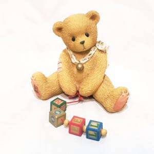 May include: A tan-colored teddy bear figurine with a white patterned scarf and a gold bell necklace. The bear is holding a red string attached to colorful alphabet blocks. The blocks spell out the word "NOEL".