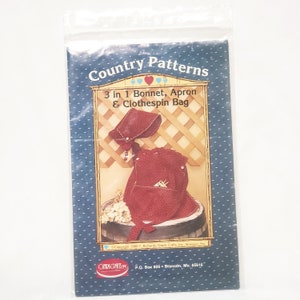 May include: A sewing pattern for a bonnet, apron, and clothespin bag. The pattern is printed on white paper with a blue background and features a red and white gingham pattern. The pattern is titled "Country Patterns 3 in 1 Bonnet, Apron & Clothespin Bag".
