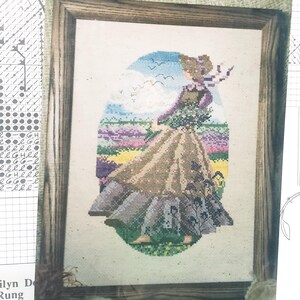 May include: A framed cross-stitch embroidery depicting a figure in a long, tan dress and bonnet standing in a field of purple and yellow flowers. The scene includes a blue sky with white clouds and birds, all rendered in detailed, colorful thread stitches.