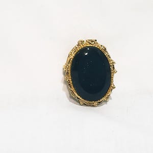 Puede incluir: Un anillo dorado con una piedra preciosa ovalada de color verde oscuro. La piedra está engastada en un marco dorado ornamentado. Una joya clásica, ideal para ocasiones especiales.