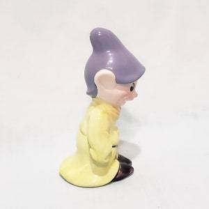May include: A ceramic figurine of a dwarf from the Disney movie Snow White and the Seven Dwarfs. The dwarf is wearing a yellow coat and a purple hat. He is looking down and has a grumpy expression.