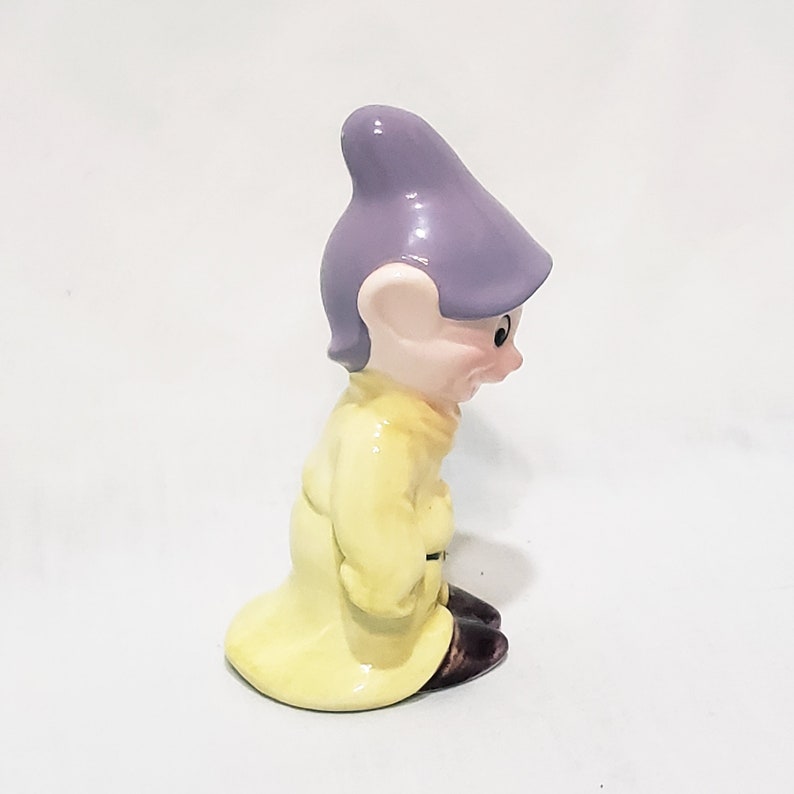 May include: A ceramic figurine of a dwarf from the Disney movie Snow White and the Seven Dwarfs. The dwarf is wearing a yellow coat and a purple hat. He is looking down and has a grumpy expression.