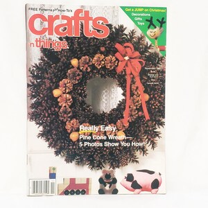 Puede incluir: Una portada de revista con una corona de piñas y un lazo rojo. La revista se titula "crafts 'n things" con el texto "Really Easy Pine Cone Wreath - 5 Photos Show You How!" y decoraciones navideñas.