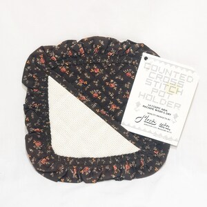 May include: A brown and floral cross stitch pot holder with a white Aida cloth insert. The holder has a ruffled edge and a tag that reads "COUNTED CROSS STITCH POT HOLDER".