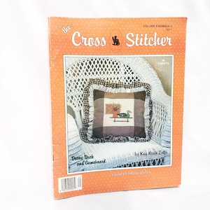 Puede incluir: La revista The Cross Stitcher, volumen 4, número 5, presenta un patrón de punto de cruz contado para una almohada con un diseño de pato señuelo y tablero de juego. La portada de la revista muestra la almohada en una silla de mimbre blanca.