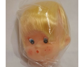 May include: A vintage doll head with blonde hair, blue eyes, and rosy cheeks, packaged in a clear plastic bag. The doll head has a painted face with freckles and a small red mouth. The doll head is a collectible item.