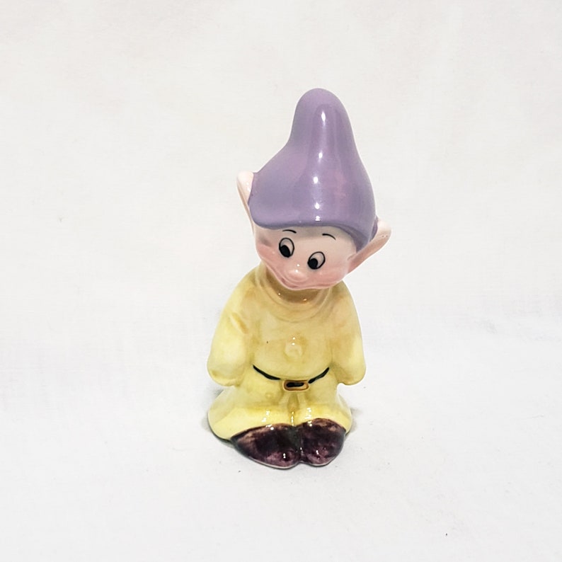 May include: A small ceramic figurine of a dwarf from the Disney movie Snow White and the Seven Dwarfs. The dwarf is wearing a yellow shirt, a purple hat, and brown shoes. He is looking down.