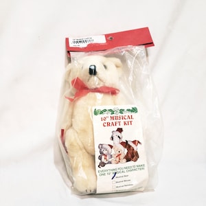 May include: A cream-colored teddy bear with a red ribbon, packaged in a clear plastic bag. The package includes a label that reads "10" Musical Craft Kit" and features illustrations of musical characters. The bear is approximately 10 inches in size.