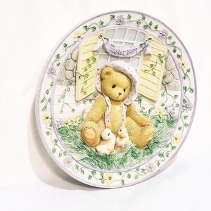 Könnte beinhalten: Dekorative Osterplatte mit einem Teddybären mit Mütze, der mit zwei Entenküken in einer Gartenszene sitzt. Die Platte hat einen floralen Rand und ein Banner mit der Aufschrift "Easter 1997". Die Farben sind Gelb, Grün und Weiß.
