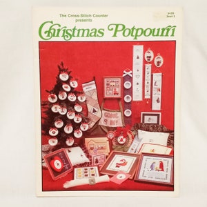 Pode incluir: Uma capa de livro com fundo vermelho e o título "Christmas Potpourri" em letras verdes. A capa apresenta uma variedade de padrões de ponto cruz, incluindo uma meia de Natal, uma árvore, enfeites e uma figura do Papai Noel.