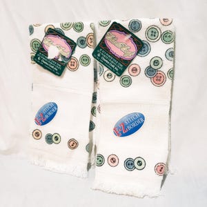 May include: Two white hand towels with a button pattern in pastel colors. Each towel has a fringed edge and a stitched border. Tags with the text "Charles Craft" are attached.