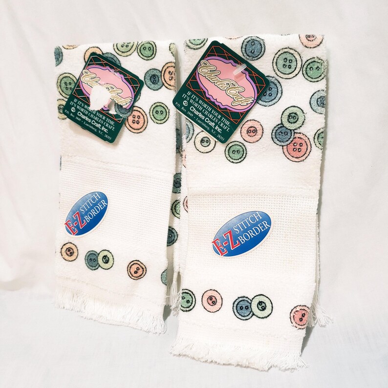 May include: Two white hand towels with a button pattern in pastel colors. Each towel has a fringed edge and a stitched border. Tags with the text "Charles Craft" are attached.