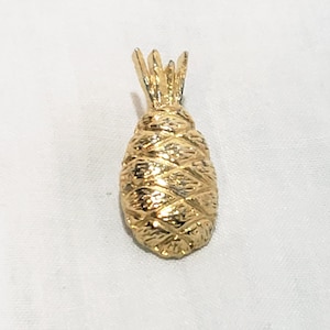 Op de afbeelding: Een goudkleurige metalen hanger in de vorm van een ananas. Het stuk heeft een getextureerd, kruislings gearceerd oppervlak op het lichaam om de schil van de vrucht na te bootsen en een puntig, bladvormig bovendeel. Het ontwerp is solide.