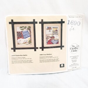 May include: Two embroidery kits, one with a clothesline and quilts, the other with a cozy kitchen scene. Both kits are in brown frames and include 14-count Aida cloth, cotton embroidery floss, a die-cut mat, a tapestry needle, a graph, and instructions. The Creative Circle logo is in the bottom right corner.