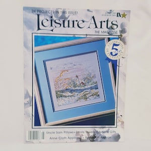 Puede incluir: Portada de la revista Leisure Arts con un diseño de punto de cruz de un faro en una playa nevada. La revista celebra 5 años de excelencia. La portada también presenta una lista de proyectos, que incluyen almohadas Uncle Sam, toallas Estate, regalos adorables, aplicaciones de tela Anne y afganos estadounidenses.