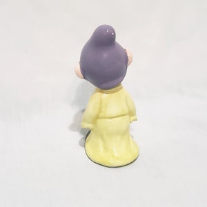 May include: A small ceramic figurine of a character wearing a yellow robe and a purple hat. The figurine is standing on a white surface.