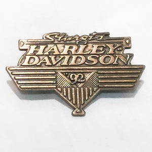May include: A gold-colored metal pin with the words "Sturgis Harley-Davidson" and the number "92" in the center.