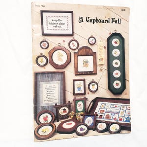 May include: A craft pattern book titled A Cupboard Full featuring various cross-stitch designs. The cover displays multiple framed embroidery projects including small circular frames, rectangular wooden frames, and a long vertical green frame.
