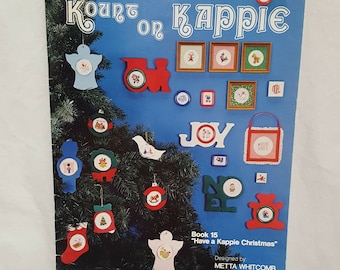 May include: A book titled "Kount on Kappie" with a Christmas theme. The cover features a Christmas tree with ornaments and a variety of cross stitch designs. The book is for counted cross stitch and needlepoint. The book is by Kappie Originals Ltd. and is from 1980.