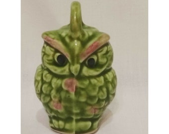 May include: A green ceramic owl figurine with pink accents and black eyes. The owl has a textured surface and a small loop on its head.