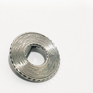 May include: A silver circular brooch with a textured, concentric circle design. The brooch has a small opening in the center.