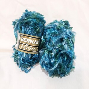 May include: Two skeins of Bernat Boa yarn in shades of teal, blue, and green. The yarn has a fluffy, textured appearance, with the brand label visible on one skein. Ideal for knitting or crochet projects.