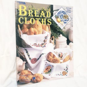 Puede incluir: Un libro de instrucciones de Leisure Arts titulado Bake-a-Batch Bread Cloths que presenta 16 diseños de punto de cruz. La portada muestra telas blancas bordadas con patrones coloridos, incluyendo un gallo, un girasol y una colmena, sobre cestas con pan.