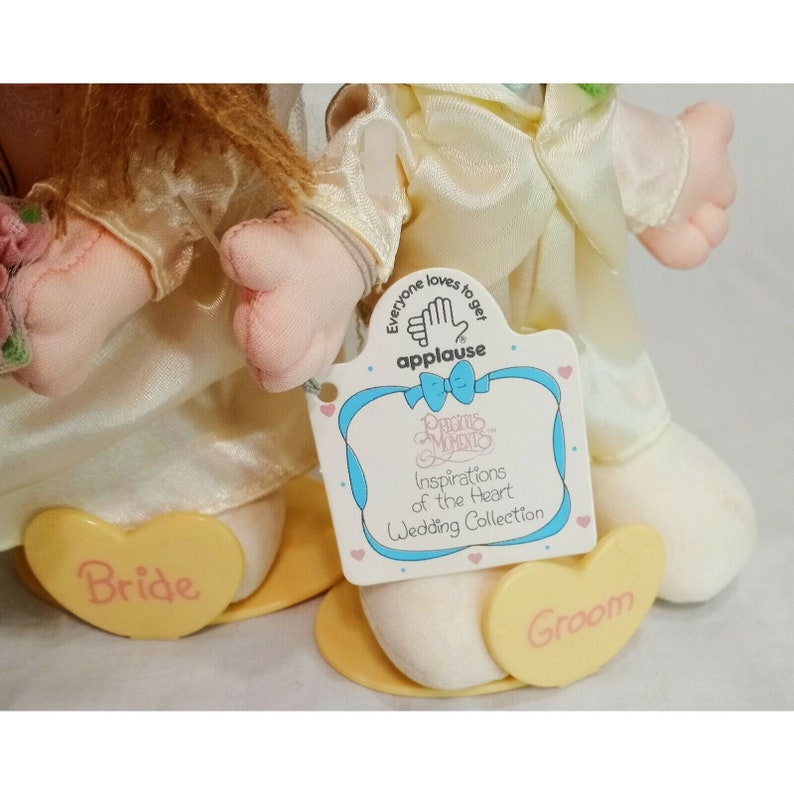 May include: A pair of plush dolls dressed as a bride and groom. The bride is wearing a white dress with a floral embellishment and a yellow heart that says "Bride". The groom is wearing a white suit with a yellow heart that says "Groom". The dolls are part of the Precious Moments Inspirations of the Heart Wedding Collection.