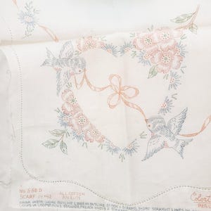 May include: A white cotton fabric panel for embroidery featuring a printed design of two birds holding a ribbon that forms a heart shape surrounded by flowers. The text reads No 538 D SCARF 14x42 ALL COTTON RN 21179 EMBR. INSTR. WORK OUTLINE LINES.