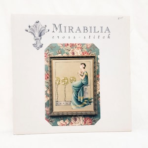 May include: A Mirabilia cross-stitch pattern featuring a framed image of a woman in a teal gown holding flowers. The image is set against a floral and teal background, with the brand name at the top.