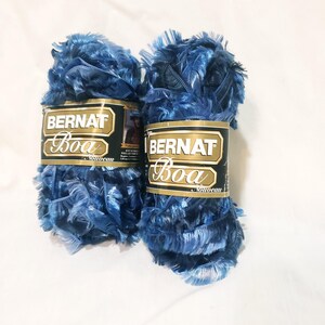 May include: Two spools of Bernat Boa Nouveau yarn in shades of blue. The yarn has a fluffy, feathery texture. Each spool has a label with the brand name and product details. Ideal for knitting and crochet projects.