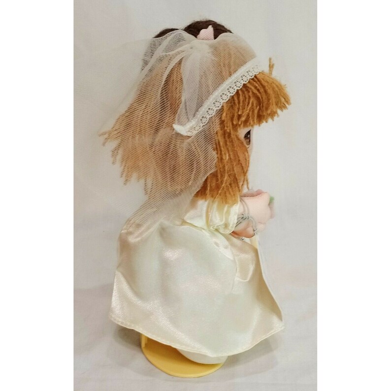 May include: A white bridal doll with brown yarn hair, wearing a white veil and holding a bouquet of flowers. The doll is standing on a yellow base.