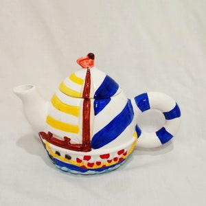 May include: A white ceramic teapot shaped like a sailboat with blue, yellow, and red stripes. The teapot has a lid and a handle shaped like a life preserver.