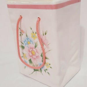 Może przedstawiać: Biały ceramiczny wazon w kształcie torby na zakupy z różowymi uchwytami i różową wstążką wokół górnej części. Wazon ma kwiatowy wzór z różowymi, niebieskimi i żółtymi kwiatami.