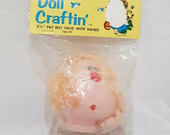 May include: A plastic doll head with blonde hair and a pink face. The head is 3 1/2 inches tall and has hands attached. The packaging says "Doll Craftin' T.M. 3 1/2" FAT BOY HEAD WITH HANDS 161-91"