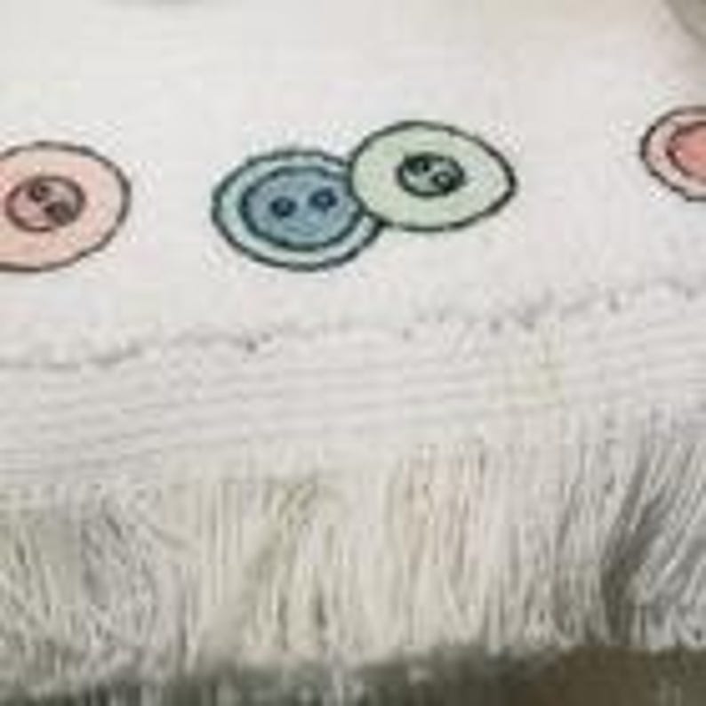 May include: White fringed towel with embroidered pastel-colored buttons. The buttons are in shades of pink, blue, and green, each with a black stitched outline and two small black dots in the center, adding a touch of whimsy to the design.