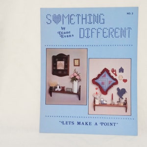 Puede incluir: Un libro de manualidades titulado "Something Different" de Diane Evans. La portada del libro presenta dos imágenes de un estante con una variedad de artículos hechos a mano, incluyendo una almohada, un gallo y una pieza enmarcada de punto de cruz. El libro es parte de una serie llamada "Lets Make a Point".