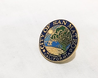 Op de afbeelding: Een ronde, goudkleurige pin met een blauwe rand. De pin toont de woorden "CITY OF SAN MATEO INCORPORATED CALIFORNIA". Het midden toont een landschap met bomen, water en een zon.