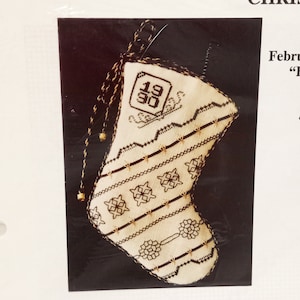 May include: A cream-colored Christmas stocking with a detailed cross-stitch design in brown. The stocking features a geometric pattern, the year "1990", and a hanging loop with gold-colored beads. The overall theme is holiday decor.