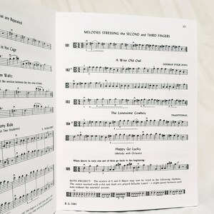 May include: A page from a music instruction book featuring printed musical notation for string instruments. The page includes exercises titled Melodies Stressing the Second and Third Fingers, A Wise Old Owl, The Lonesome Cowboy, and Happy Go Lucky.